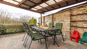 Un patio al aire libre con muro de ladrillo, una mesa con sillas y una zona cubierta con techo metálico.