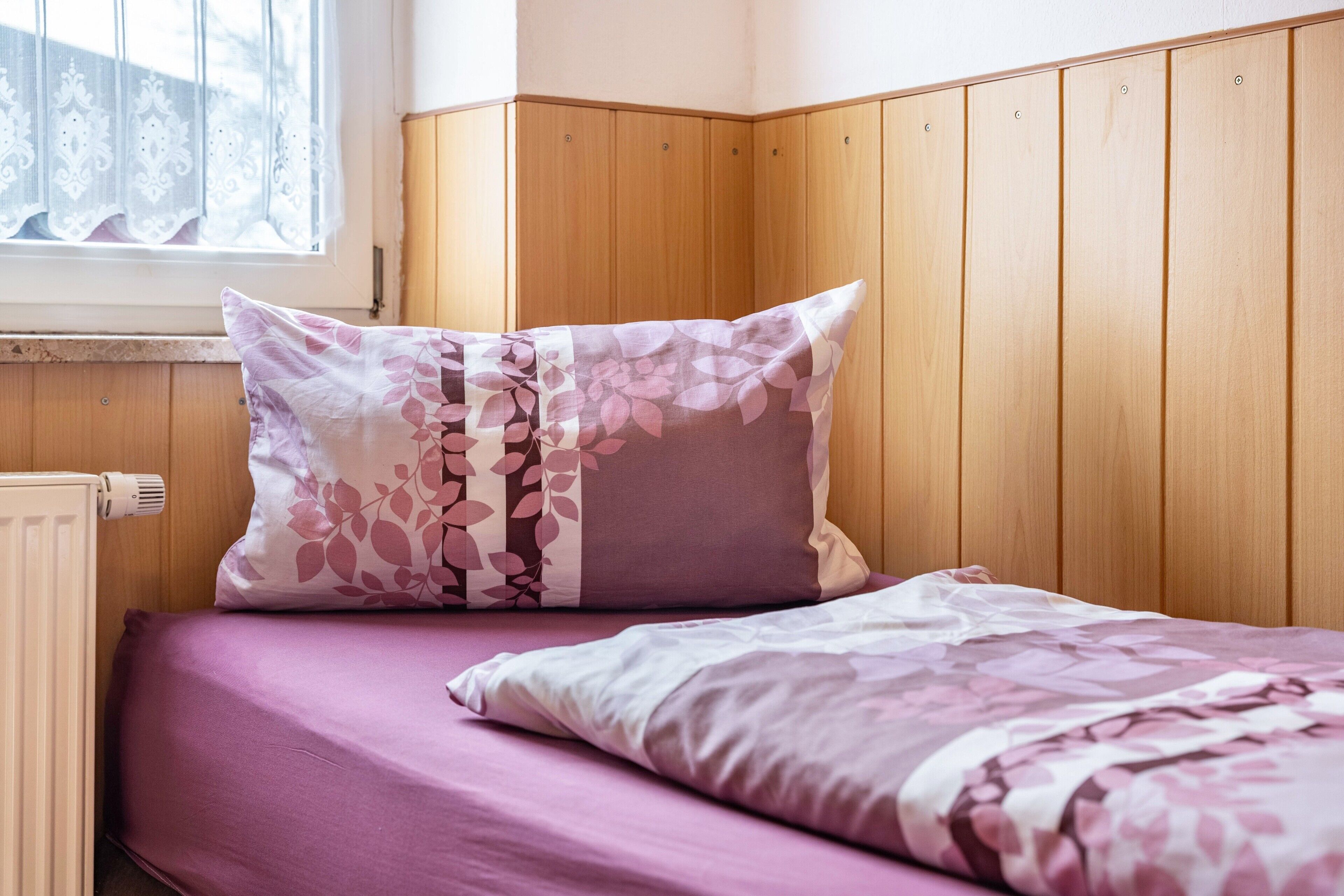 Quarto com uma cama, janela com cortinas e painéis de madeira nas paredes.
