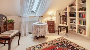 Una habitación con un ventanal grande, una mesa redonda con mantel de encaje, una estantería, una silla y una alfombra estampada.