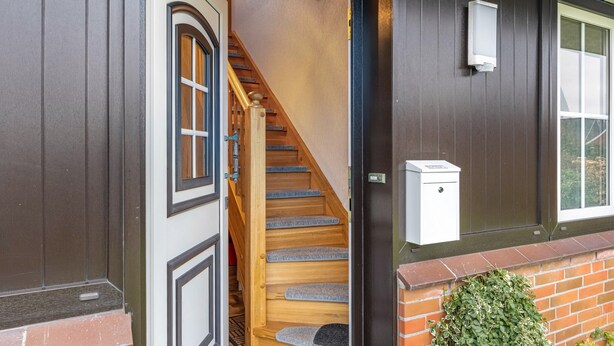Una escalera que lleva a una puerta de madera, un cuadro eléctrico blanco en la pared, y un muro de ladrillo con una pequeña planta verde.