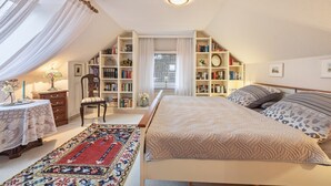 Un dormitorio con cama, estantería, silla, mesita de noche con lámpara y ventana con cortinas.