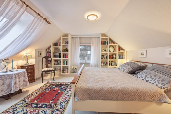 Une chambre à coucher avec un lit, une bibliothèque, une chaise, une table avec une lampe et une fenêtre avec des rideaux.