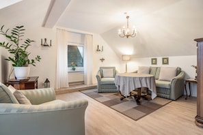 Une pièce chaleureuse de style loft, avec un plafond haut, un lustre, une table à manger, deux fauteuils, un canapé, une plante en pot et une vue sur l’extérieur.
