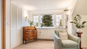 Una habitación con un radiador, un aparador de madera, un sillón verde claro, una planta en maceta y una ventana con vistas a un árbol.