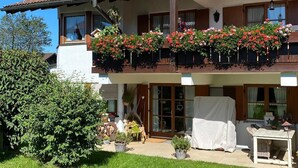 A two-story building with a balcony adorned with red flowers, a covered outdoor seating area, and a well-maintained garden.