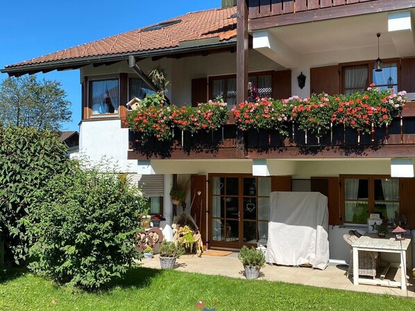 A two-story building with a balcony adorned with red flowers, a covered outdoor seating area, and a well-maintained garden.