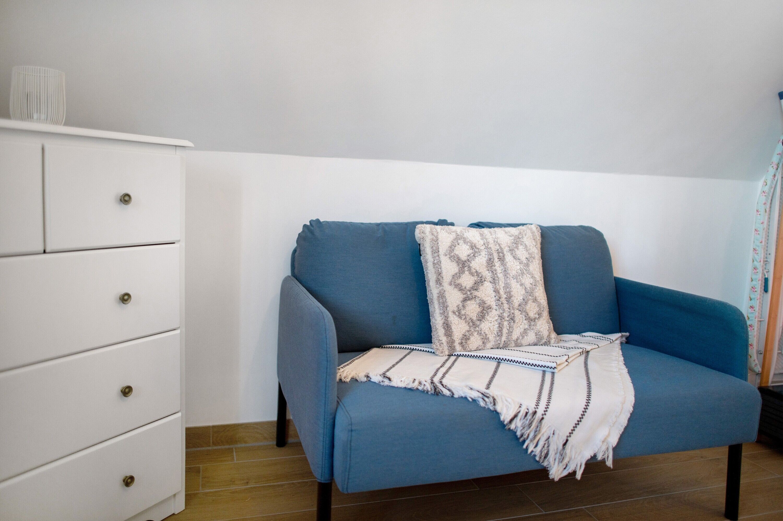 A room with a blue sofa, a white dresser with drawers, and a patterned throw blanket.