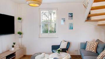Een moderne woonkamer met een flatscreen televisie, een houten dressoir, een blauwe bank, een klein rond tafeltje en een raam met uitzicht op bomen.