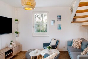 Ett modernt vardagsrum med en platt-TV, en träkonsol, en blå soffa, ett litet runt bord och ett fönster med utsikt över träd.