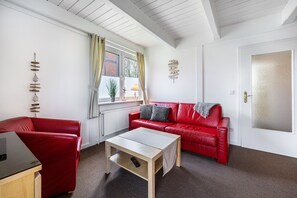 Une pièce comprenant un canapé en cuir rouge, une table basse en bois, un fauteuil rouge, une fenêtre avec des rideaux et une porte dotée d’un panneau en verre dépoli.
