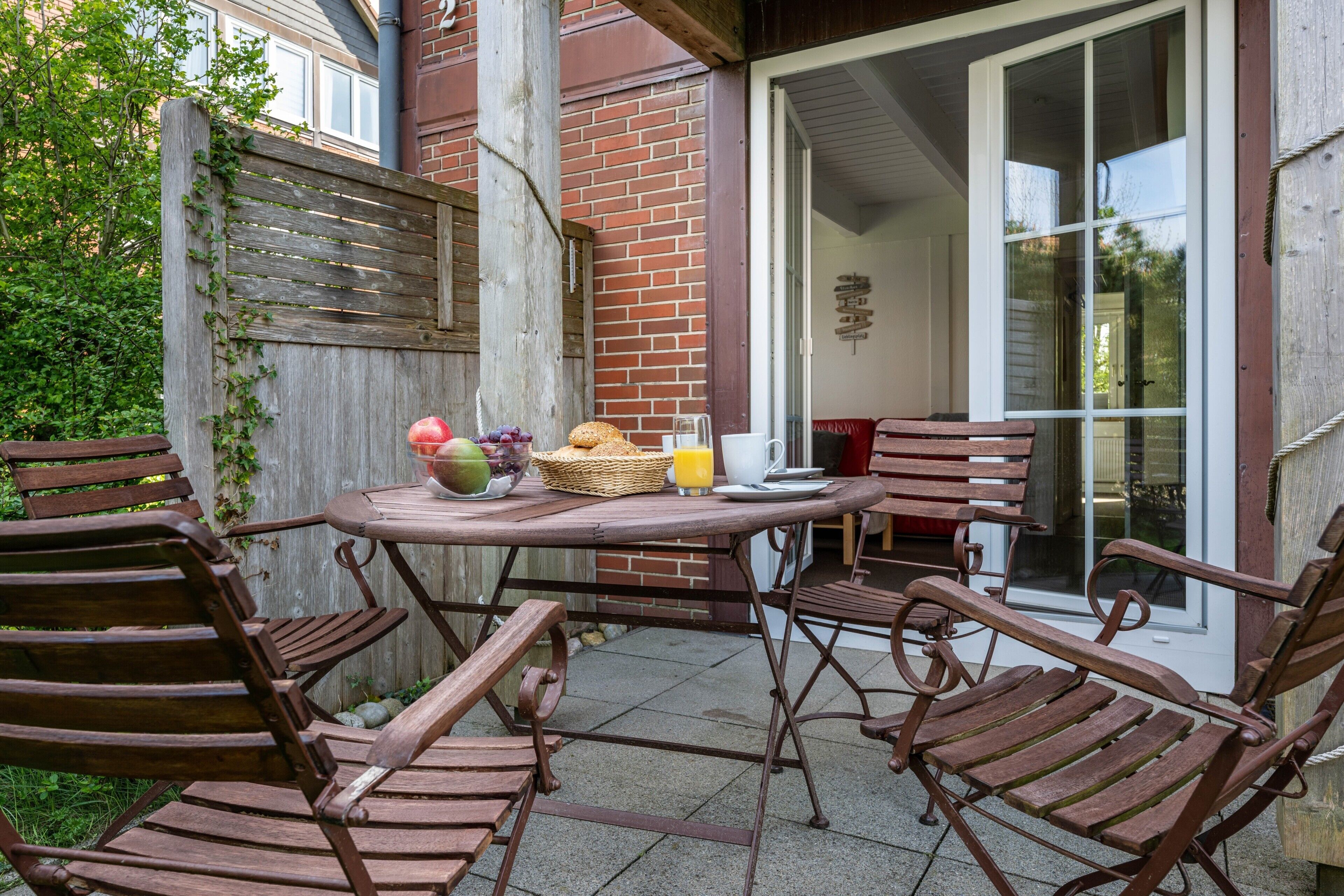 A patio with a table and chairs, a basket with fruits and a glass of juice, and a brick wall.