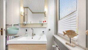 A bathroom with a white sink, a mirror, a shelf with toiletries, and a window with blinds.