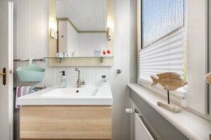 Une salle de bain avec un lavabo blanc, un miroir, une étagère contenant des articles de toilette et une fenêtre avec des stores.