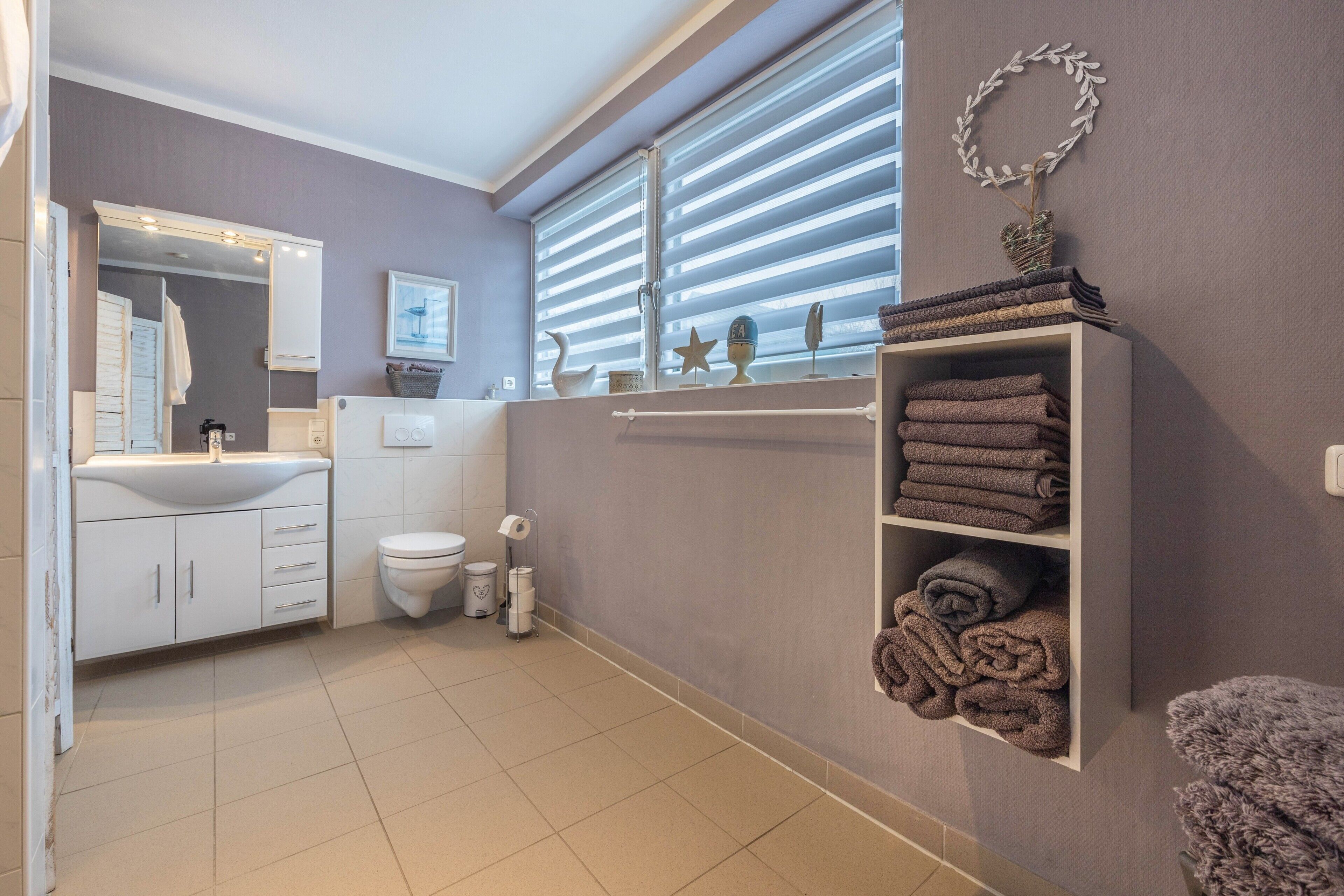 Une salle de bain moderne avec un grand miroir, des meubles blancs, des toilettes et une étagère avec des serviettes.