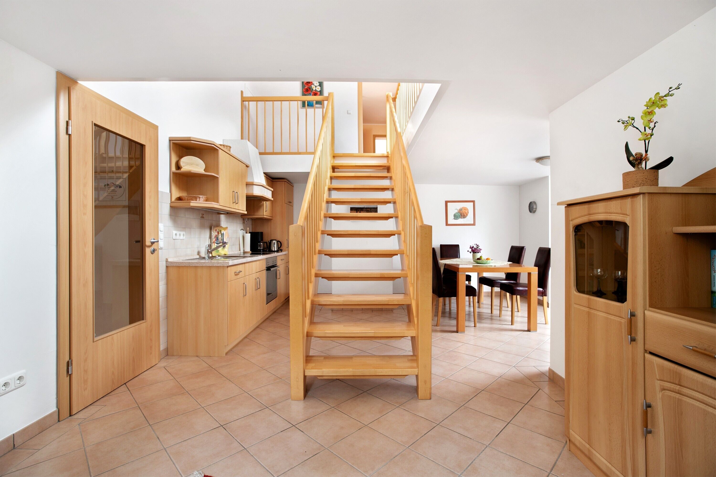 A modern kitchen with wooden cabinets, a staircase, and a dining area with chairs and a table.