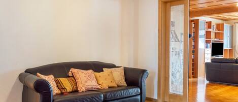A living room with a black leather sofa, patterned cushions, a wooden floor, and a view into a dining area.