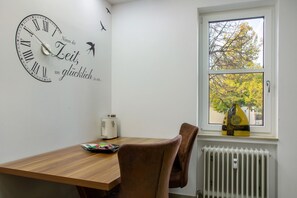 Una habitación con una mesa de madera, un reloj con texto en alemán, un radiador y una ventana con vistas a árboles.