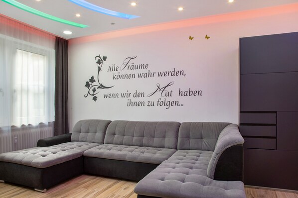 Una sala de estar moderna con un sofá modular, un muro decorativo con una cita, y un ventanal con cortinas translúcidas.