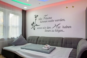 Et moderne hotelværelse med en grå sofa, en pænt redt seng og en væg med et motiverende citat på tysk.
