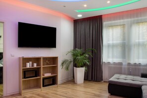 Una sala moderna con televisor de pantalla plana, estanterías de madera, una planta en maceta, un sofá gris y una ventana con cortinas.
