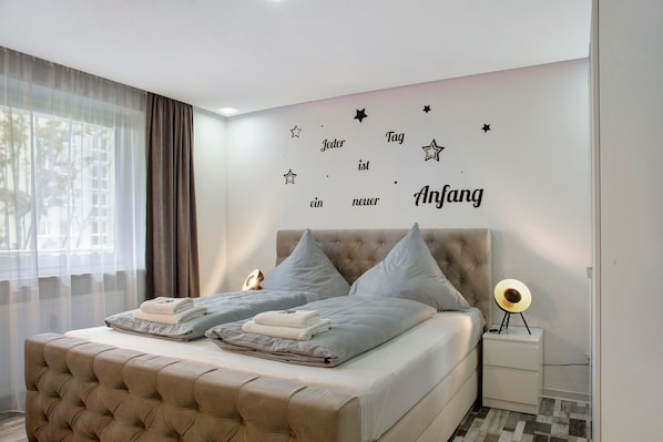 A bedroom with a tufted headboard, two pillows, a bedside table with a lamp, and a wall decal with text and stars.