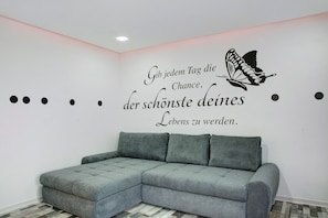 A room with a grey sectional sofa, a wall with a motivational quote and a butterfly decal, and a patterned floor.