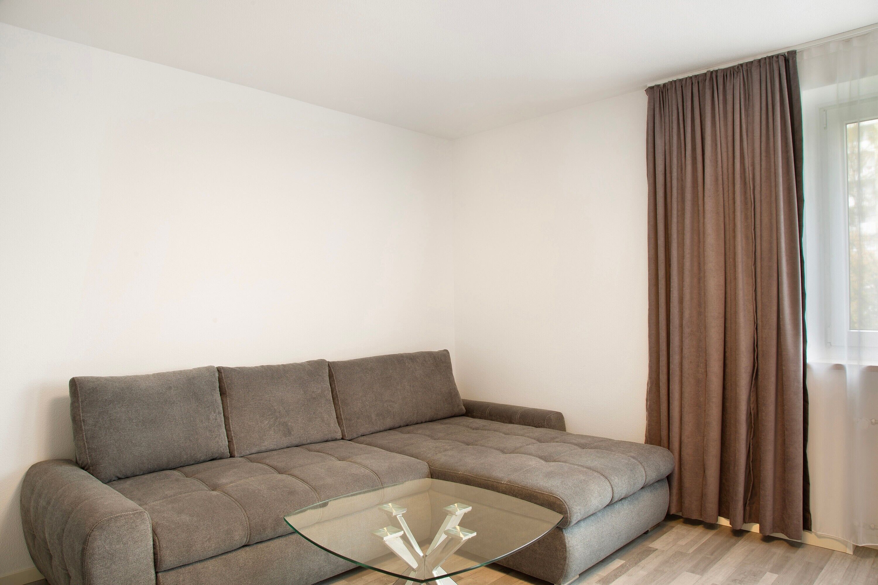 A living room with a grey sofa, a glass coffee table, and a window with brown curtains.