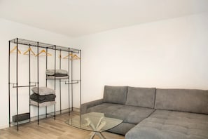 A modern living room with a grey sofa, a glass coffee table, and a metal wire shelf with clothes.