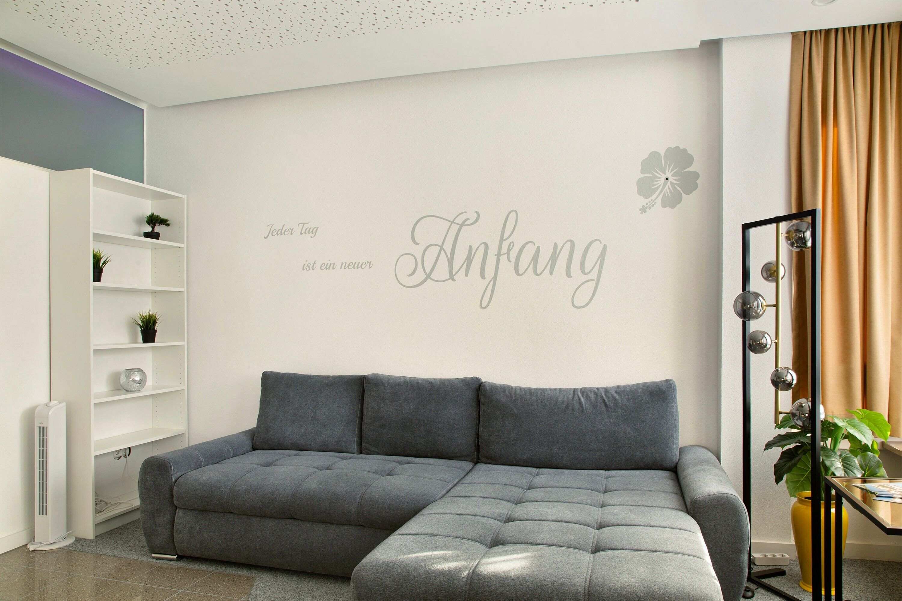 Una sala de estar moderna con un sofá seccional gris, una estantería blanca con plantas, un espejo montado en la pared y una pared decorativa con texto y una flor.