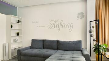 Una sala de estar moderna con un sofá seccional gris, una estantería blanca con plantas, un espejo montado en la pared y una pared decorativa con texto y una flor.