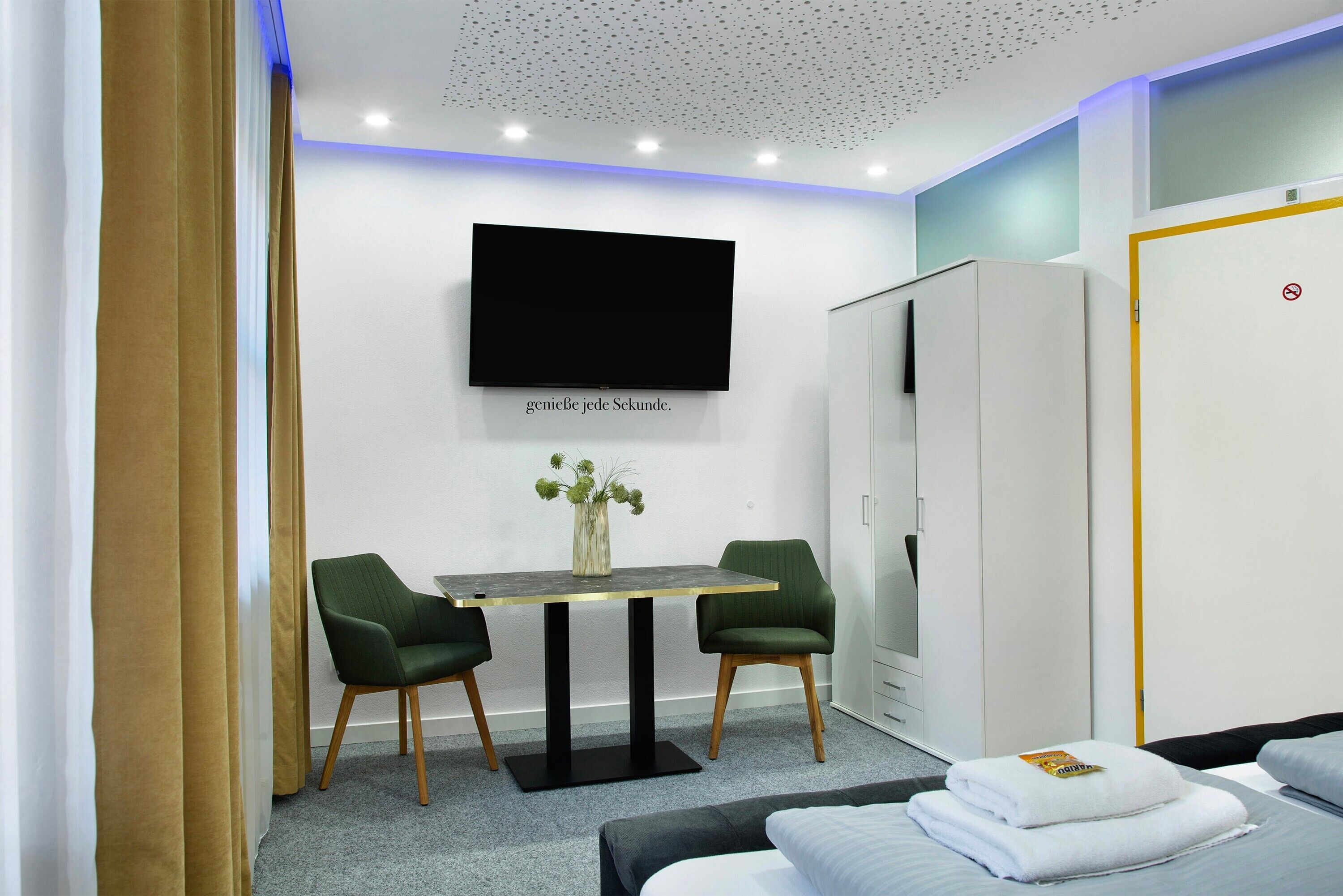 Une chambre d’hôtel moderne équipée d’une télévision à écran plat, d’une petite table avec des chaises, d’une armoire et d’un lit avec des serviettes soigneusement pliées.