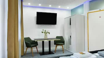 A modern hotel room with a flat-screen TV, a small table with chairs, a wardrobe, and a bed with neatly folded towels.