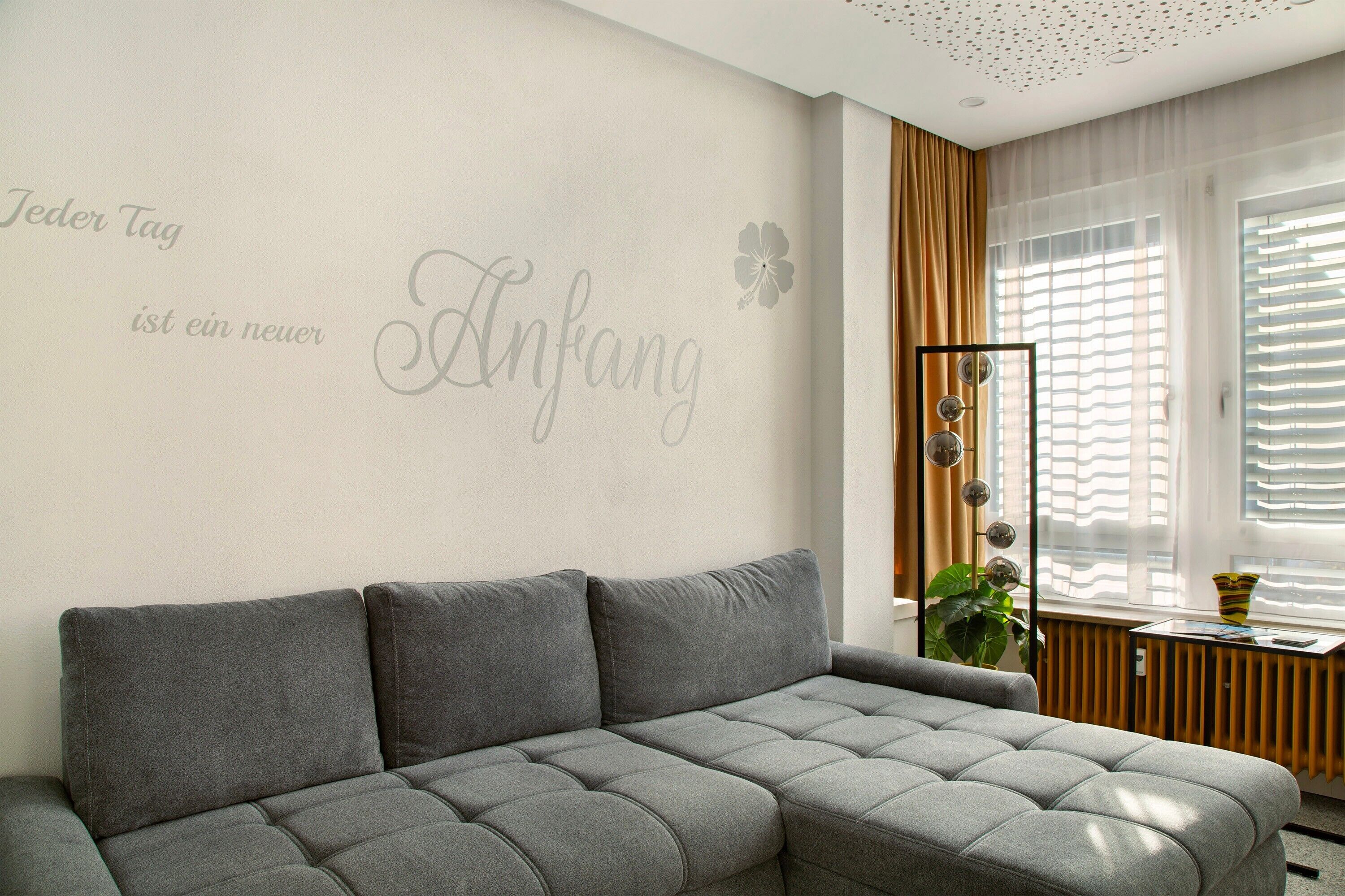 Un salon moderne avec un canapé gris, un mur orné d’un motif floral et une fenêtre avec des stores.
