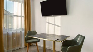 A hotel room with a flat-screen TV mounted on the wall, a table with two chairs, a window with sheer curtains, and a radiator. - Apartment 'Neuburg 1 - New' with Wi-Fi (Neuburg an der Donau)