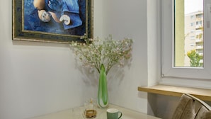 Ein Zimmer mit einem Gemälde, einer Blumenvase, einem Tisch mit einer Tasse und einem Fenster mit Blick auf Gebäude.