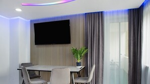 Una habitación moderna con una televisión de pantalla plana montada en la pared, una mesa de comedor con cuatro sillas, una planta en maceta y una puerta corredera de vidrio.