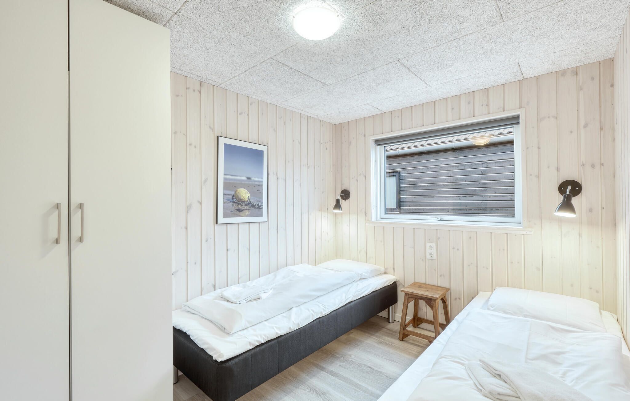 A room with two beds, a wardrobe, a window with blinds, a picture on the wall, and a small stool.
