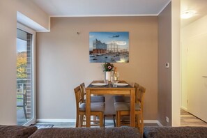 A dining area with a table set for two, a painting above the table, and a view of the outdoors through a window. - Apartment 'Ferienwohnung Neu Wulmstorf' with Balcony (Neu Wulmstorf)