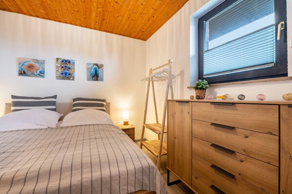 A bedroom with a bed, wooden dresser, and a window with blinds. - Apartment 'Ferienwohnung 27 Sandbank, 80 Qm' with Sea View, Balcony and Wi-Fi (Wangerland)