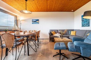 Un salon moderne avec un plafond en bois, un coin repas avec des chaises en osier, un canapé gris et un fauteuil bleu.