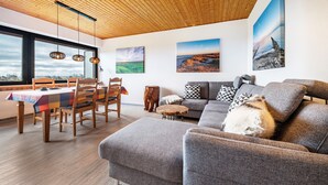 Un salon moderne avec un plafond en bois, un canapé gris, une table à manger avec des chaises et une vue dégagée grâce à de grandes fenêtres.