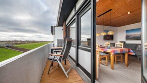 Un balcon donnant sur un champ verdoyant, un coin repas avec une table et des chaises, et un intérieur moderne avec de grandes fenêtres et un plafond en bois.
