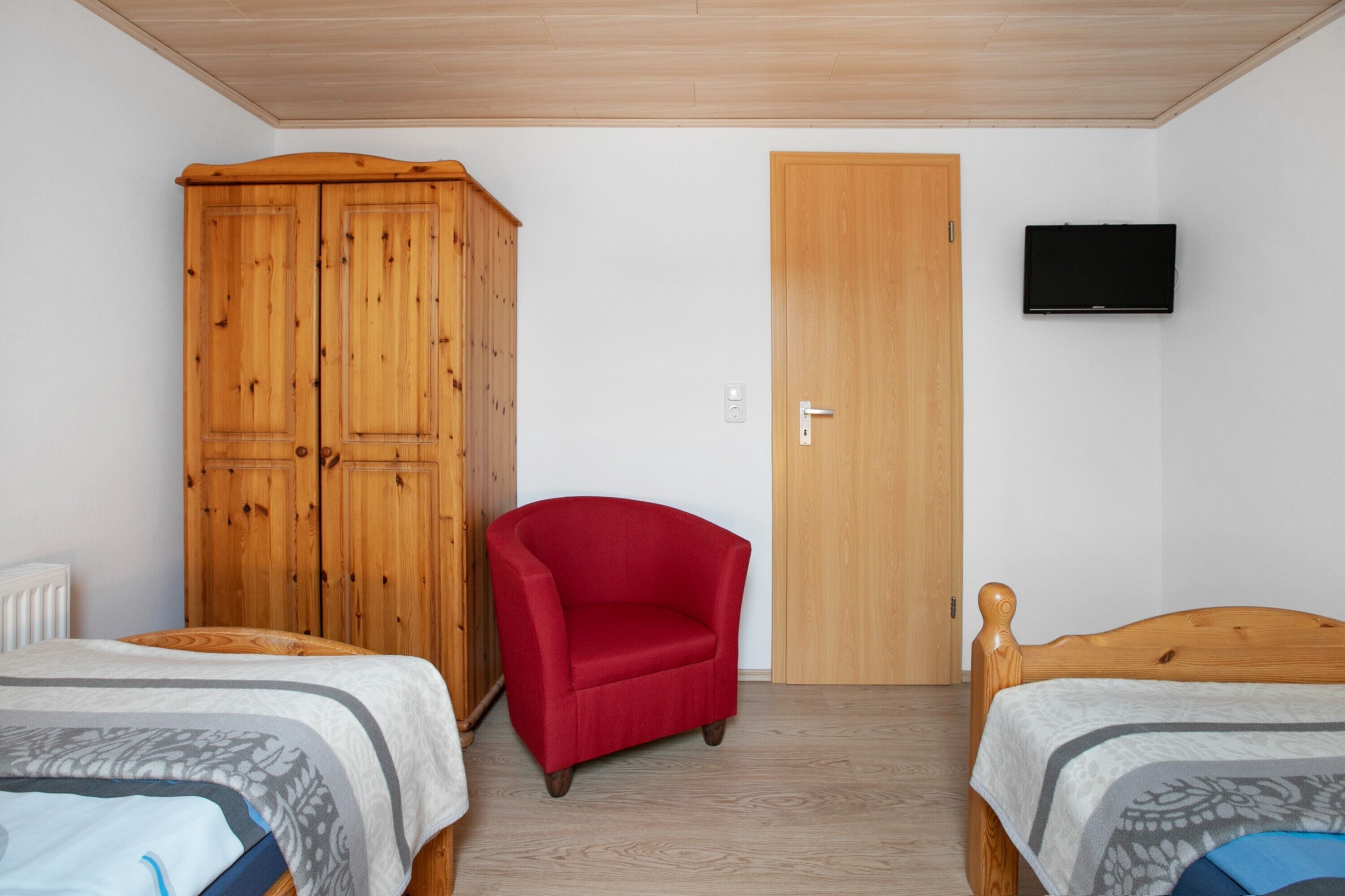 Ein Schlafzimmer mit zwei Betten, einem roten Sessel, einem Holzschrank, einer Holztür und einem Fernseher an der Wand.