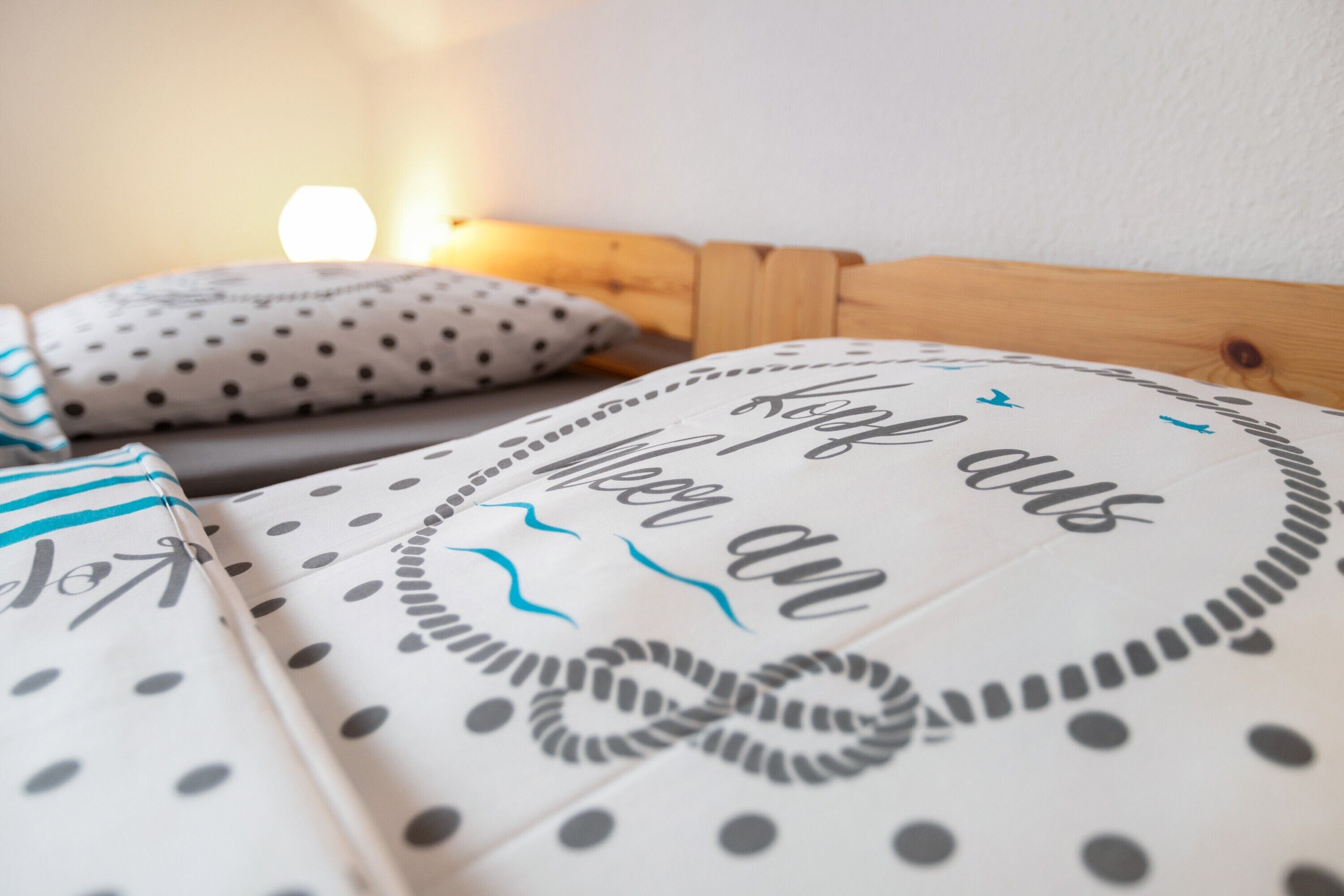 En seng med prikket og stribet sengetøj, en pude med lignende mønster og en vægmonteret lampe.