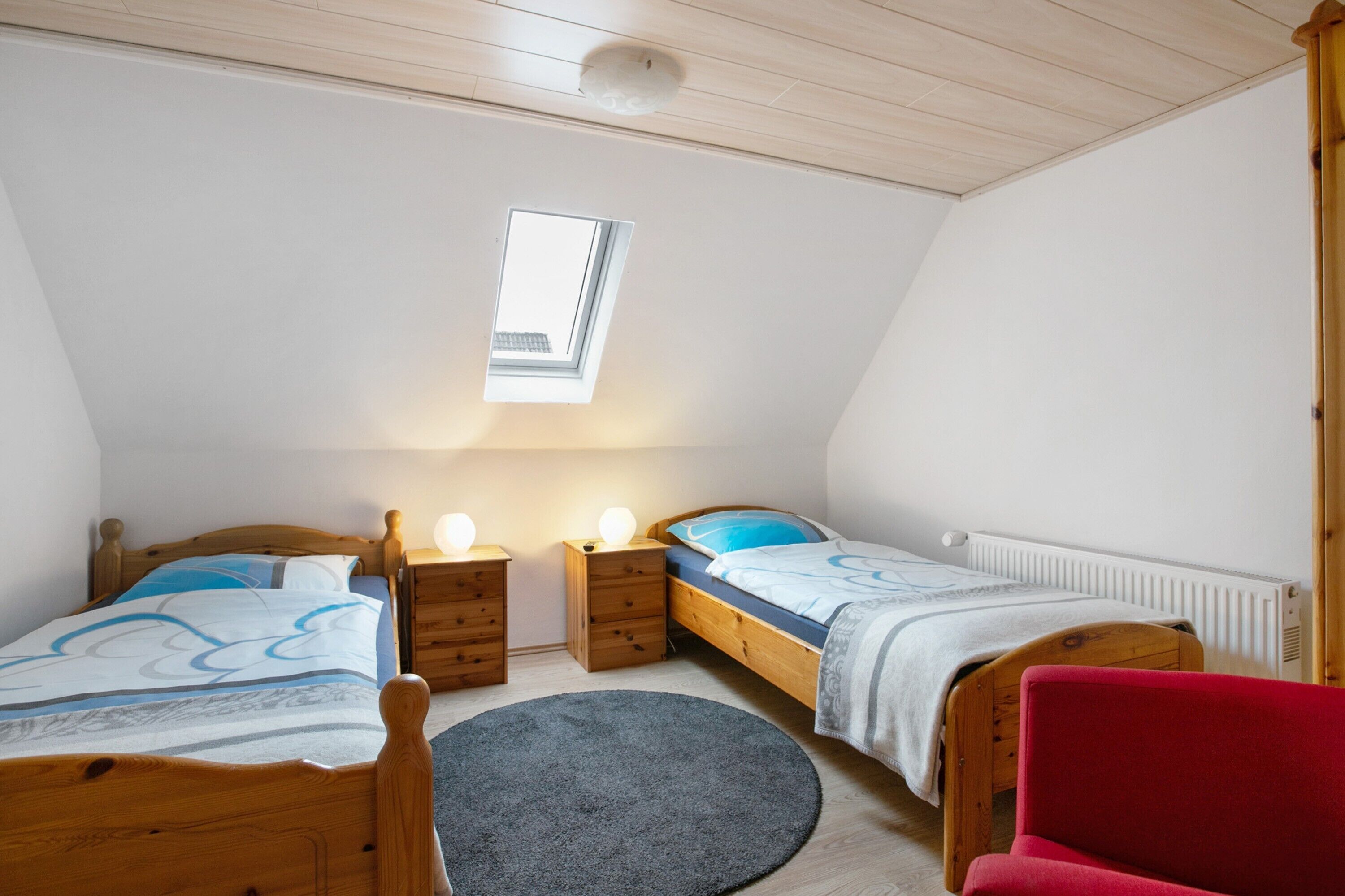 Un dormitorio con dos camas de madera, un tragaluz pequeño, una silla roja y una alfombra redonda.