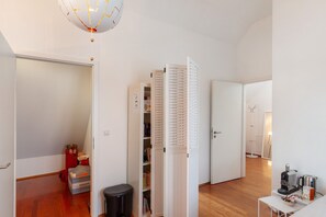 Une pièce avec un plancher en bois, une porte blanche, une bibliothèque, une petite table avec une bouilloire et un luminaire au plafond.