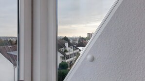 Une vue à travers une fenêtre donnant sur un quartier résidentiel avec de nombreux immeubles, un ciel dégagé et un mur blanc orné d’une petite protubérance ronde.