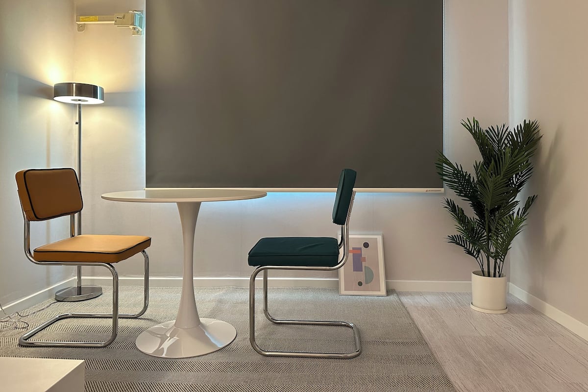 Ein Konferenzraum mit einem weißen Tisch, zwei Stühlen, einer Projektorleinwand, einer Stehlampe und einem Blumentopf.