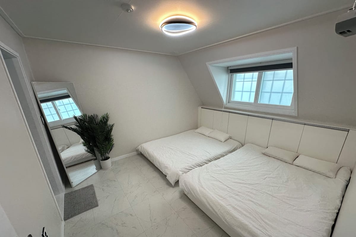Ein Schlafzimmer mit zwei Betten, einer Zimmerpflanze, einem Fenster und einer Deckenleuchte.