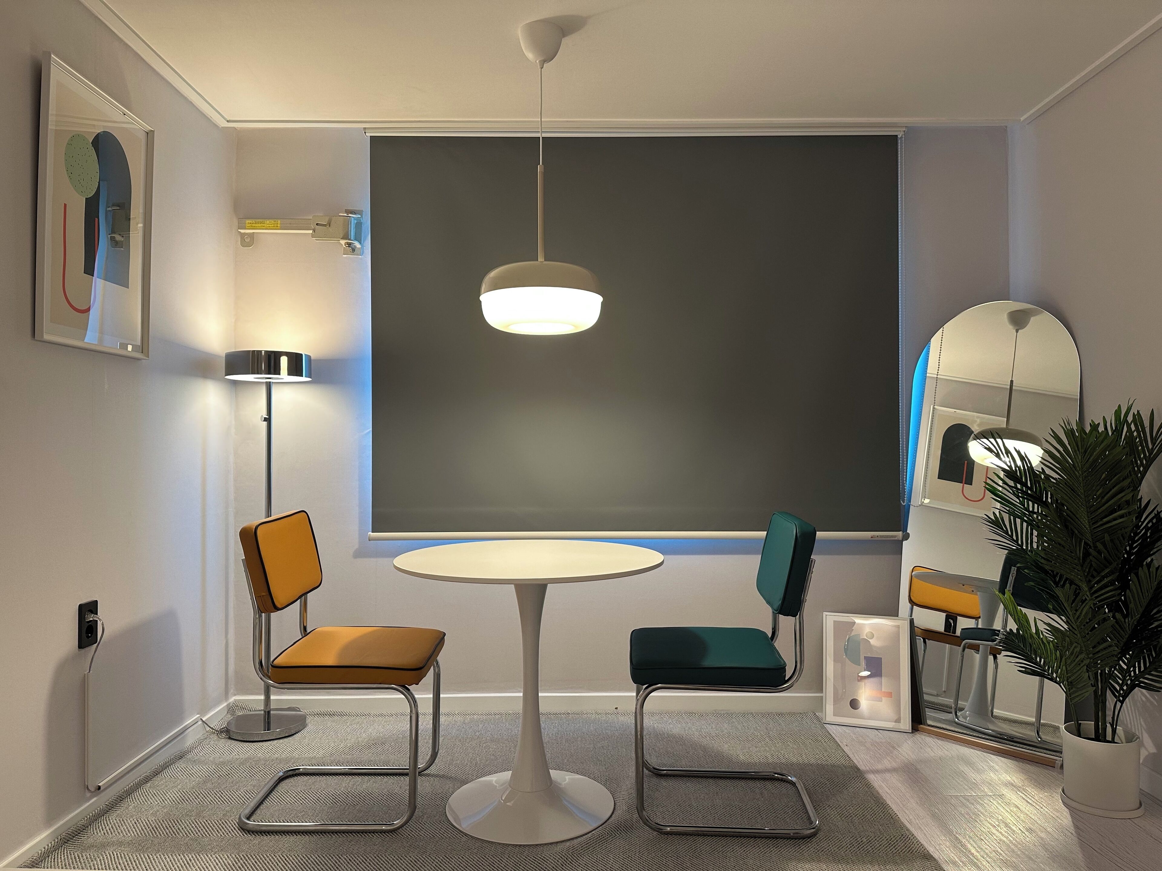 Ein modernes Zimmer mit rundem Tisch, zwei Stühlen, einer Stehlampe, Deckenleuchte, einem großen Fenster und einem Blumentopf.